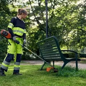 a woman wearing high-visibility workwear, using the Husqvarna 525iLXT Trimmer to cut grass underneath a park bench