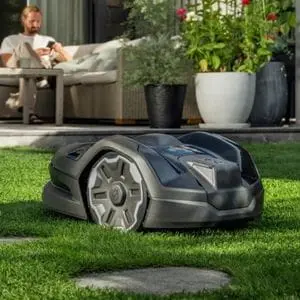 the Husqvarna Automower® 305E NERA Robotic Mower mowing a lawn, with a man sat on patio furniture in the background