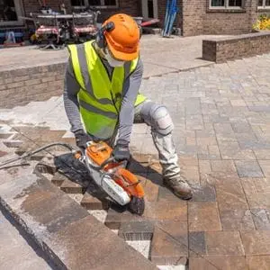 a man wearing a hardhat, ear defenders and a dust mask, using the STIHL TSA 300 Cordless Cut-off Machine to cut small setts on a driveway