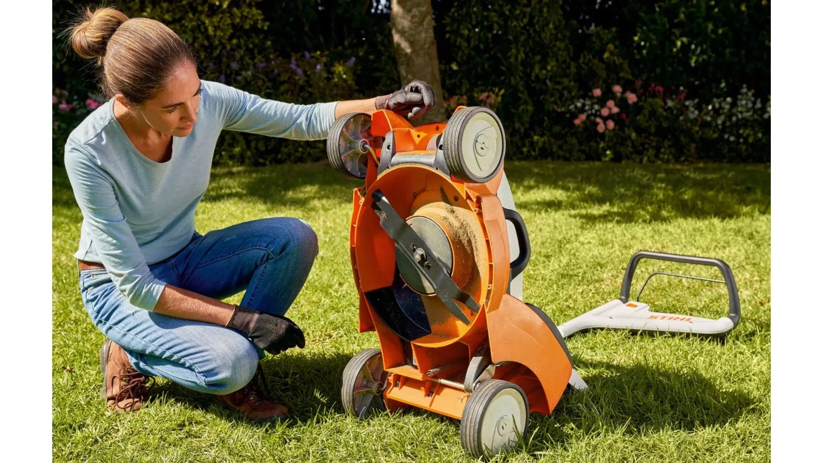 a woman with her hair in a bun, kneeling down to examine the underside of an orange STIHL lawn mower
