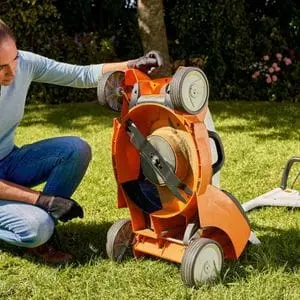 a woman knelt on one knee, examining the underside of a STIHL lawn mower