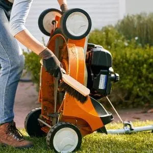 a woman cleaning an orange lawnmower's cutting deck using a brush
