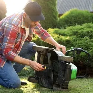 a man wearing a red checked shirt and blue baseball cap, removing a lawnmower blade using a screwdriver