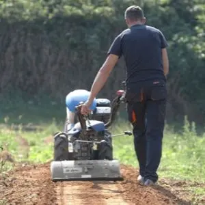 a rearview shot of a man using the BCS 780HY PowerSafe® Hydrostatic Petrol Two Wheel Tractor with Rotary Hoe attachment to prepare soil in a field