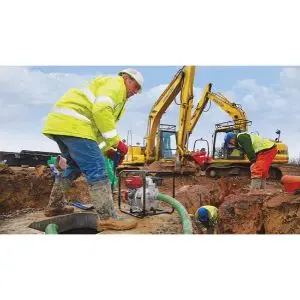 Construction workers using a Honda petrol water pump to drain a flooded ditch, with a yellow digger operating in the background.