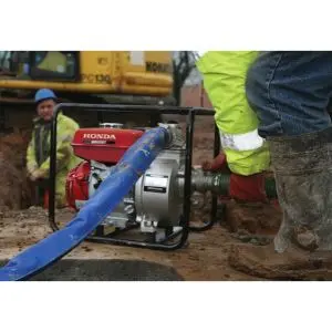 A construction worker connecting a green suction hose to a Honda petrol water pump, with a blue discharge hose attached and another worker standing in an excavation in the background.