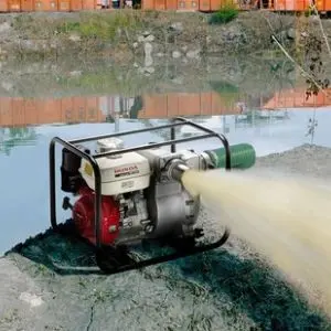 Honda petrol trash pump positioned beside a muddy pond, discharging a powerful stream of water through its outlet, with a green suction hose connected to the pump.