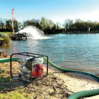 Honda petrol trash pump positioned on a sandy lakeshore, with a green suction hose extending into the water and a large fountain spraying in the background.