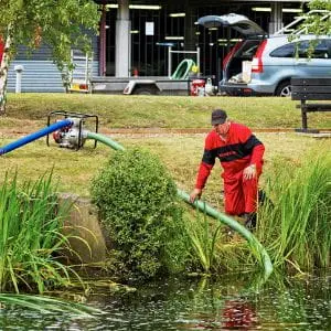 A man in red overalls positioning the green suction hose of a Honda petrol water pump in a garden pond, with the pump on the bank behind him.
