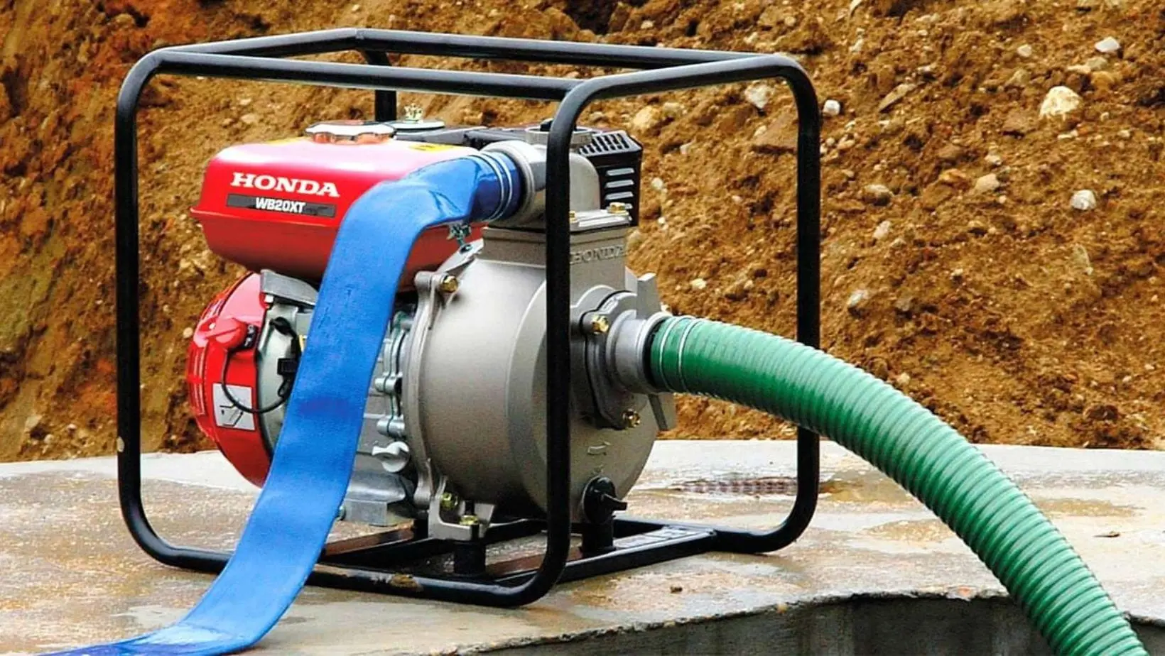 Honda WB20XT petrol water pump with blue discharge hose and green suction hose, positioned beside an excavation on a construction site.
