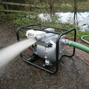 Honda WT20 petrol trash pump operating beside a muddy pond, discharging a powerful stream of water, with a green suction hose connected to the inlet.