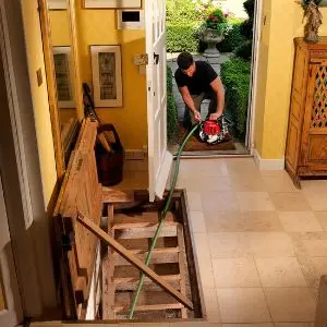A man operating a portable Honda petrol water pump outside a house, with a green hose extending into an exposed indoor basement.