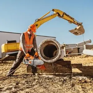 a man wearing an orange hardhat and high-visibility vest, using the STIHL TS 440 Petrol Cut-off Machine to cut a section of precast concrete on a building site, in front of a yellow crane