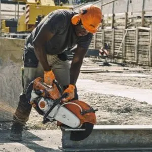 A construction worker wearing an orange hard hat, ear defenders, and protective gloves uses a STIHL TS 500i Petrol Cut-Off Machine to cut a concrete slab on a building site.