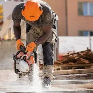 A construction worker wearing an orange hard hat, ear defenders, and protective gloves uses a STIHL petrol disc cutter to cut concrete on a building site, with water spraying from the cutting wheel to suppress dust.